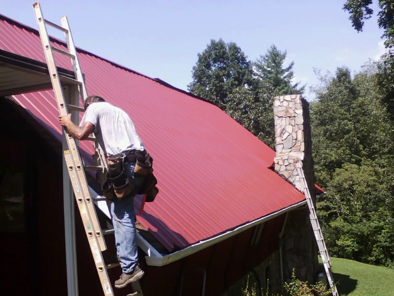 Roofer on a ladder beside a red metal roof and stone chimney — Soffit & Fascia Repair by Roofing Brothers in Douglas City